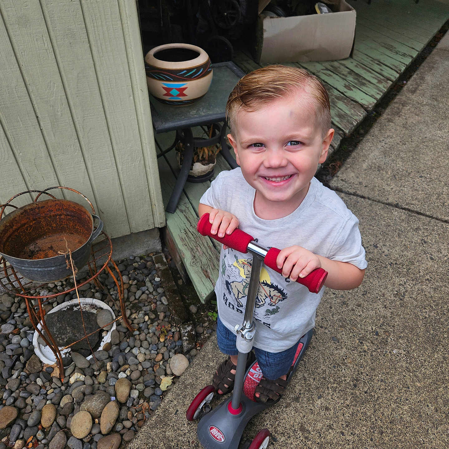 Jed joined the competition — help win amazing prizes! child, toddler, smiling, outdoor, portrait, scooter, driveway, porch, house, garden, potted_plant, rocks, pebbles, chair, pottery, playtime, blond_hair, gray_shirt, denim_shorts, happy