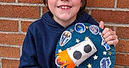 Isaac participe au concours pour gagner de l'argent avec cette photo : child, boy, smiling, brick_wall, blue_hoodie, black_pants, craft, rocket, planets, stars, space_theme, handmade, happy, portrait, standing, outdoor, casual_clothing, art_project, young, background