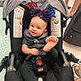 Addie is registered to the contest to win money with this photo: baby, car_seat, headband, pom_poms, smiling, foot, playful, child, indoor, luggage, floor, person, legs, toy, black_clothing, seatbelt, happy, infant, accessory, celebration