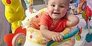 Addie joined the competition — help win amazing prizes! baby, activity_center, toy, colorful, smiling, indoor, child, plant, home, floor, foot, face, clothing, orange_shirt, plastic, seat, play, happy, cute, infant