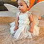 Addie is registered to the contest to win money with this photo: baby, child, costume, fairy_wings, headband, white_dress, carpet, indoor, person, sitting, cute, toddler, flower_pattern, orange_pants, furniture, living_room, soft_lighting, curly_hair, expression, playful