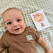 élio participe au concours pour gagner de l'argent avec cette photo : baby, infant, smiling, face, clothing, brown_shirt, milestone_card, blanket, soft_texture, cute, teddy_bear, hearts, lying_down, child, happy, portrait, indoors, person, closeup, expression