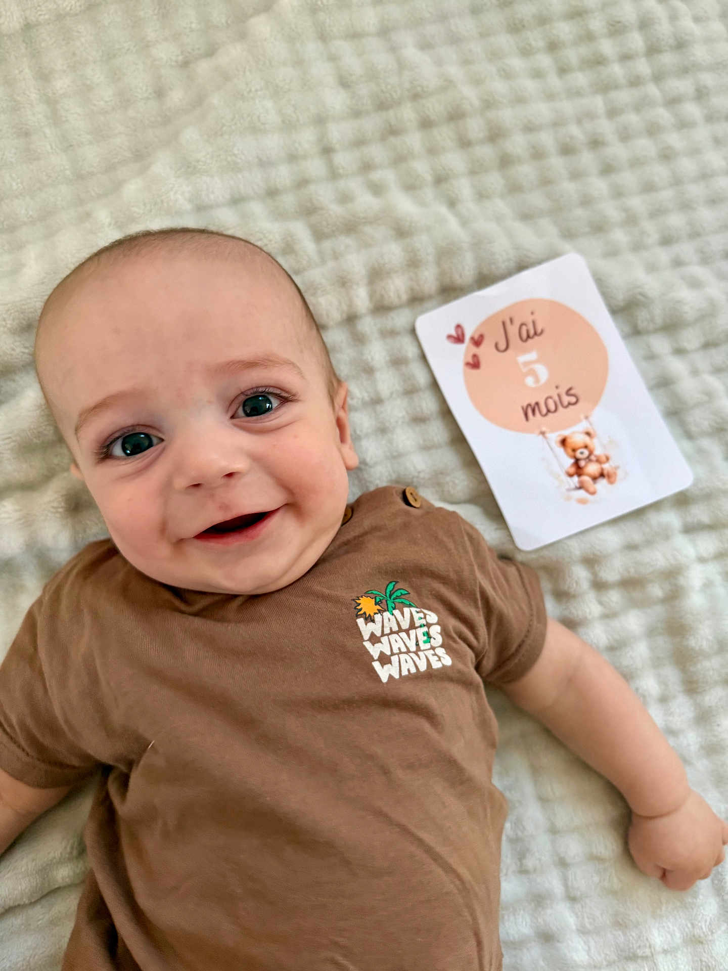 élio participe au concours pour gagner de l'argent avec cette photo : baby, infant, smiling, face, clothing, brown_shirt, milestone_card, blanket, soft_texture, cute, teddy_bear, hearts, lying_down, child, happy, portrait, indoors, person, closeup, expression