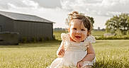 Alice is registered to the contest to win money with this photo: baby, child, infant, smiling, happy, portrait, outdoor, grass, field, countryside, barn, farm, dress, white_clothing, curly_hair, sitting, sunlight, cloudy_sky, nature, rural_landscape