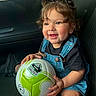 Alice is registered to the contest to win money with this photo: baby, child, toddler, smiling, happy, portrait, soccer_ball, football, sports, car, vehicle, seated, denim_overalls, black_shirt, pigtails, hair_bows, indoor, close_up, playful, joyful