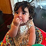 Alice is registered to the contest to win money with this photo: baby, child, toddler, sitting, eating, snack, smiling, face, curly_hair, portrait, indoor, activity_chair, colorful, floral_dress, dress, toy, playtime, home, exercise_equipment, photography