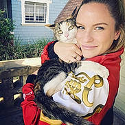 Kitty is registered to the contest to win money with this photo: animal, blue_wall, cat, daylight, embrace, face, fur, hand, house, outdoor, person, pet, plants, portrait, red_jacket, smile, tabby_cat, tail, window, woman