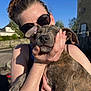woman, dog, puppy, brindle, sunglasses, smiling, hand, outdoor, sunlight, sky, happy, pet, holding, face, person, animal, nature, daytime, casual, affection