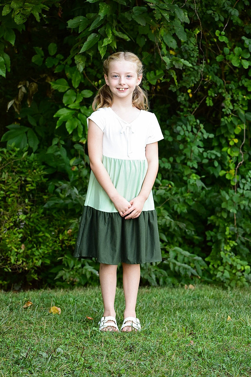 Camryn is registered to the contest to win money with this photo: brown_hair, day_dress, face, formal_wear, grass, hair, happy, jewellery, joy, knee, pattern, people_in_nature, person, plant, sandal, shrub, sleeve, smile, t_shirt, toddler