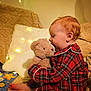 baby, teddy_bear, plaid_pajamas, red_clothing, cozy, blanket, star_pillow, fairy_lights, soft_toy, child, indoor, bedtime, cute, smiling, comfort, warm_lighting, couch, playful, portrait, infant