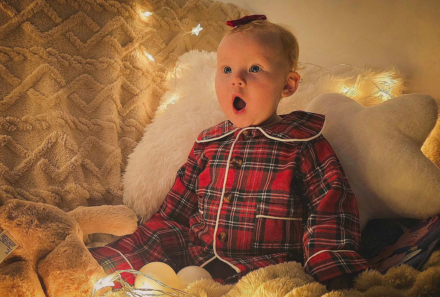 Harper is registered to the contest to win money with this photo: baby, child, pajamas, plaid, blanket, pillow, soft_toy, fairy_lights, cozy, indoor, cute, surprised, expression, warm_lighting, bedroom, holiday, decor, infant, portrait, comfortable