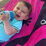 Tylio participe au concours pour gagner de l'argent avec cette photo : baby, smiling, bottle, towel, beach, sand, child, blue_clothing, happy, outdoor, sunlight, infant, person, toy, relaxing, playful, head, hand, lying_down, casual