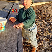 Sandro participe au concours pour gagner de l'argent avec cette photo : toddler, child, green_beanie, green_sweatshirt, jeans, brown_shoes, wooden_bench, toy_car, toy_cup, outdoor, sunlight, palm_trees, playground, daylight, face, standing, curious, expression, ground_cover, background