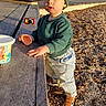 toddler, child, green_beanie, green_sweatshirt, jeans, brown_shoes, wooden_bench, toy_car, toy_cup, outdoor, sunlight, palm_trees, playground, daylight, face, standing, curious, expression, ground_cover, background