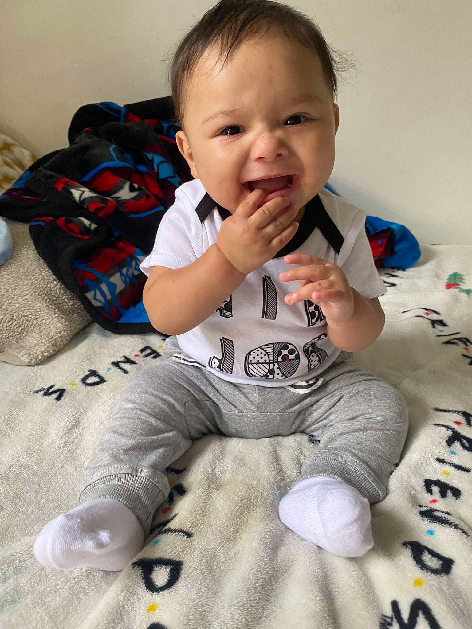 Elian is registered to the contest to win money with this photo: arm, baby, baby_toddler_clothing, cheek, child, comfort, flooring, hairstyle, happy, head, linens, long_sleeved_t_shirt, pattern, person, sitting, skin, sleeve, smile, t_shirt, thumb