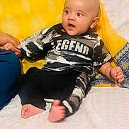 Raja Essa is registered to the contest to win money with this photo: baby, infant, child, person, clothing, black_and_white, legend, sitting, hand_holding, yellow_cushion, bed, blanket, foot, face, head, cute, expression, indoors, comfort, portrait