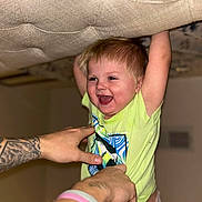 Grayson joined the competition — help win amazing prizes! toddler, child, smiling, happy, laughing, indoor, tattoo, hand, lifting, green_shirt, cushion, playful, person, baby, light_skin, short_hair, fun, joy, diaper, holding