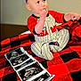 Remington joined the competition — help win amazing prizes! baby, bed, black, blanket, child, clothing, cute, face, footed_pajamas, hand, indoors, infant, person, photograph, plaid, portrait, red, sitting, striped, ultrasound