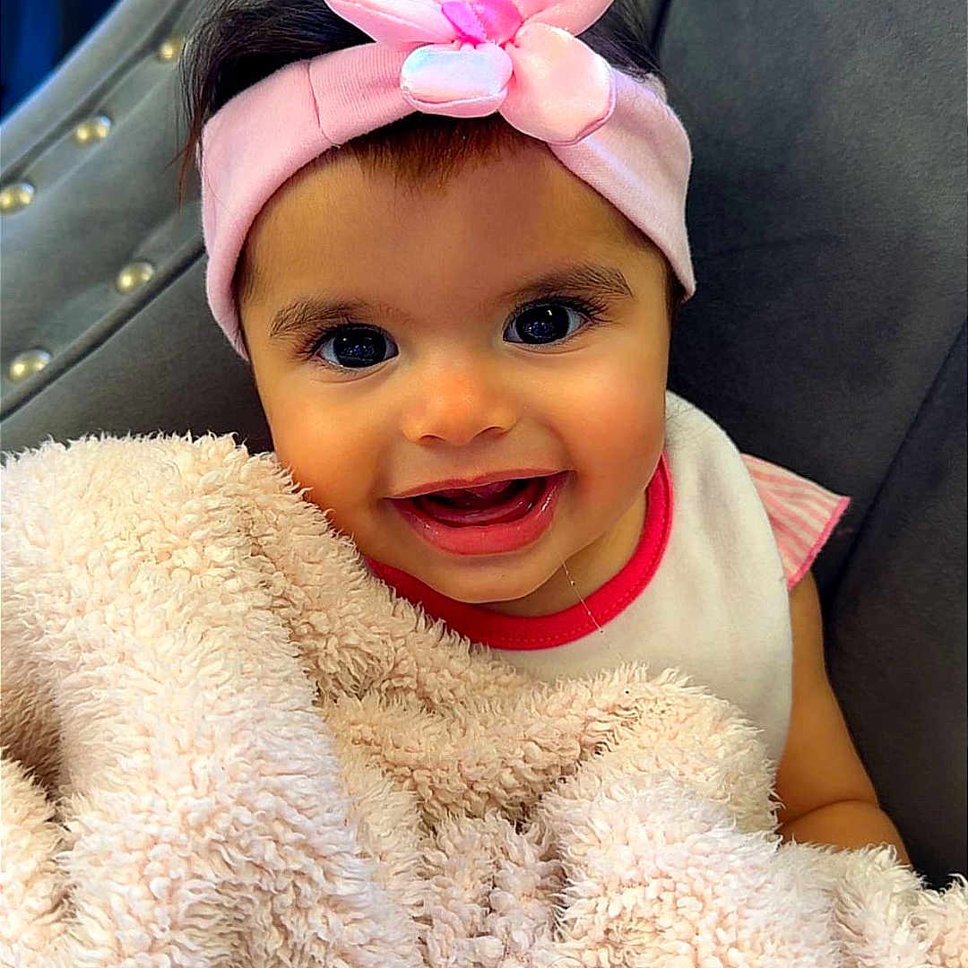 Eliana is registered to the contest to win money with this photo: baby, child, smiling, headband, pink_flower, blanket, fluffy, chair, indoor, happy, cute, person, portrait, young_child, cozy, fabric, soft, face, toddler, clothing