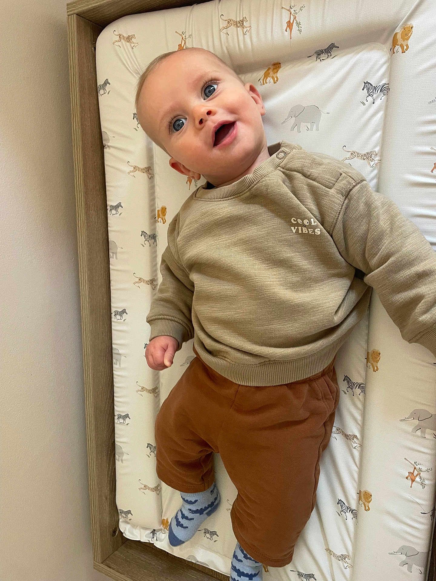 Samuel joined the competition — help win amazing prizes! baby, child, infant, smiling, blue_eyes, beige_sweater, brown_pants, patterned_socks, changing_pad, animal_print, wooden_frame, indoor, cute, happy, portrait, person, clothing, wall, laying_down, baby_bed