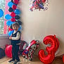 child, boy, birthday, balloons, spiderman, number_three, party, celebration, decorations, window, smile, indoor, floor, vest, sneakers, black_pants, white_shirt, happy, colorful, event
