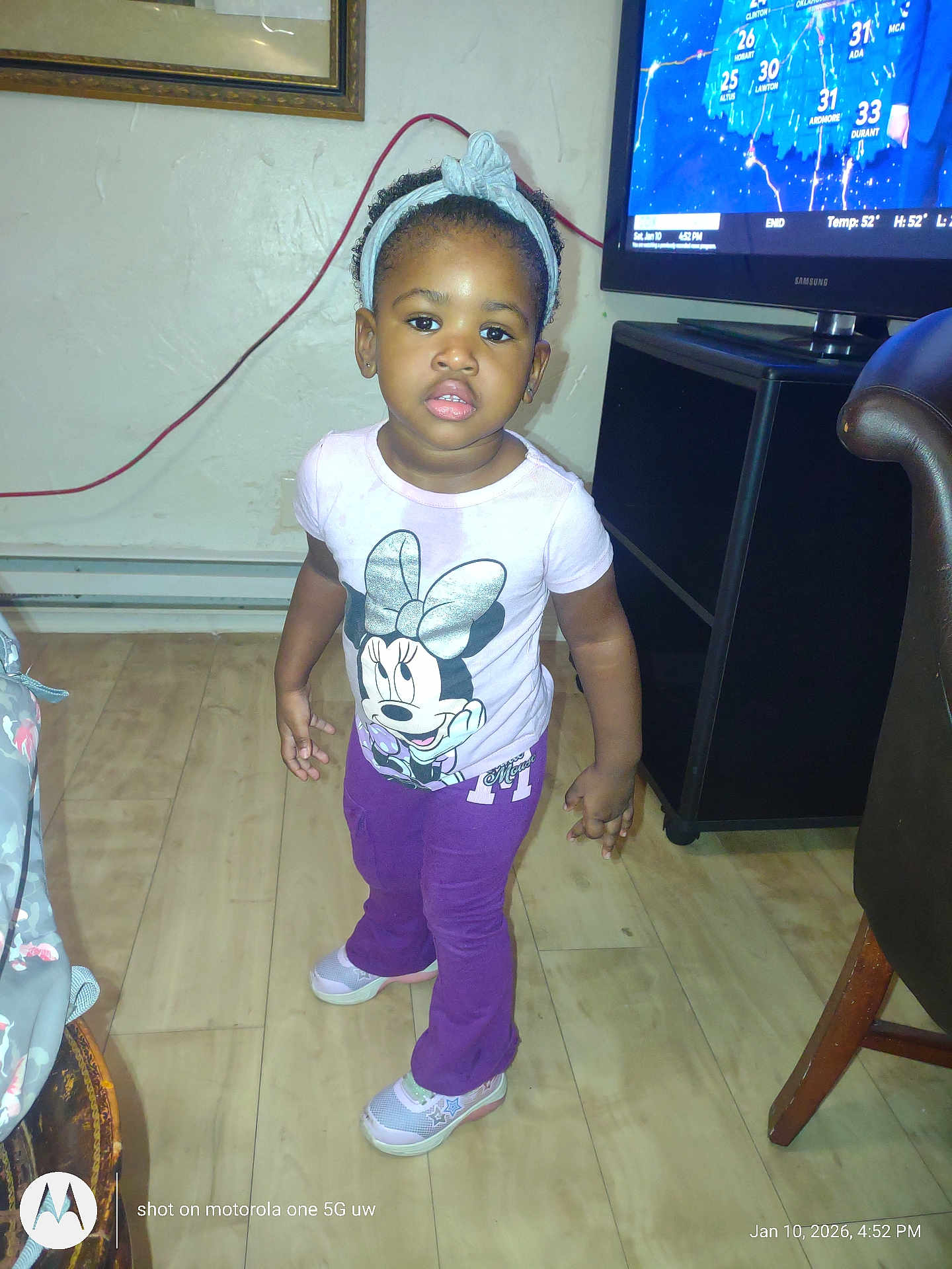 Danolia is registered to the contest to win money with this photo: toddler, child, indoor, standing, minnie_mouse, tshirt, purple_pants, sneakers, headband, wood_floor, television, weather_forecast, living_room, furniture, earrings, curly_hair, expression, person, home, casual_clothing