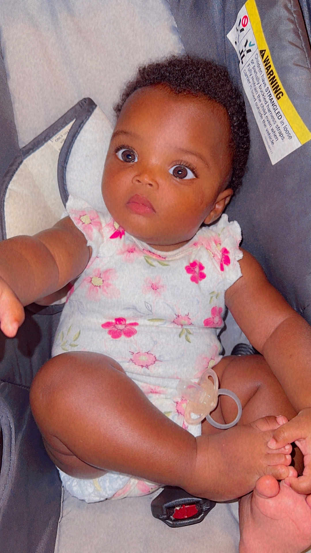 Rayne is registered to the contest to win money with this photo: baby, child, infant, car_seat, onesie, floral_pattern, foot, hand, pacifier, curious, brown_skin, indoors, seat_belt, closeup, cute, portrait, person, sitting, young, comfortable