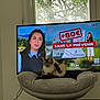 Mia participe au concours pour gagner de l'argent avec cette photo : cat, siamese_cat, television, tv_screen, news_broadcast, anchor, woman, graphic, text_on_tv, armchair, cushion, living_room, window, tree, wood_floor, floor, cable, indoor, pet, sitting