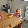 baby, child, toy, table, wooden_table, dinosaur_toy, playroom, kitchen_toy, window, radiator, floor, wall_decor, pencil_drawings, person, indoor, smiling, happy, clothing, furniture, playing