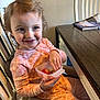 toddler, child, smiling, pajamas, high_chair, snacking, fruit, indoor, wooden_table, books, happy, face, hand, seated, furniture, table, clothing, person, breakfast, casual