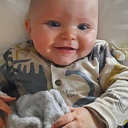 Christian-Levi is registered to the contest to win money with this photo: baby, child, smiling, blanket, bed, onesie, dinosaur, cute, infant, hand, face, comfort, sleepwear, soft, happy, indoors, portrait, young, cozy, lying_down