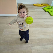 Sohan participe au concours pour gagner de l'argent avec cette photo : toddler, child, indoor, wooden_floor, green_ball, playroom, foam_mats, smiling, standing, sweater, pants, toy, hair, cute, young_child, person, flooring, casual_clothing, holding, play