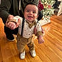 Lucca is registered to the contest to win money with this photo: baby, child, smiling, happy, standing, suspenders, bow_tie, white_shoes, adult_hand, christmas_tree, holiday, decorations, wooden_floor, indoor, person, festive, cute, celebration, home, family