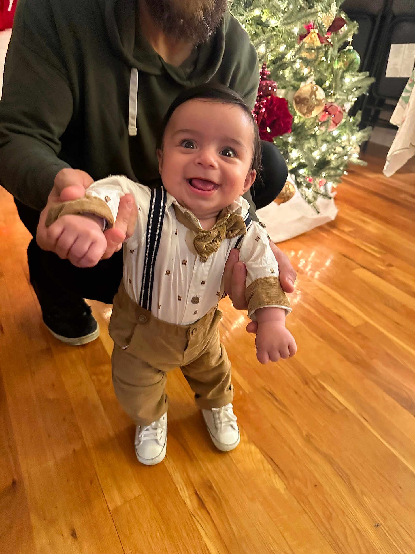 Lucca is registered to the contest to win money with this photo: baby, child, smiling, happy, standing, suspenders, bow_tie, white_shoes, adult_hand, christmas_tree, holiday, decorations, wooden_floor, indoor, person, festive, cute, celebration, home, family