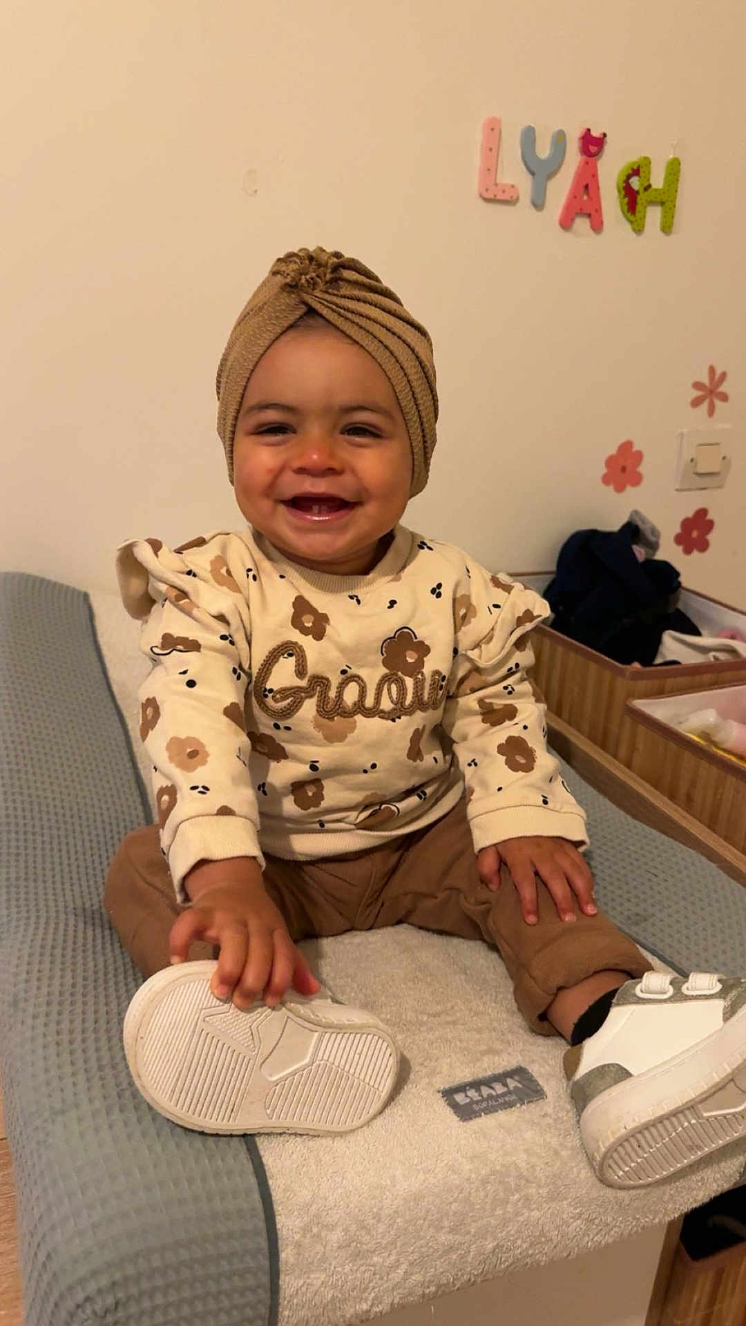 Lyah a rejoint le concours — aidez-le/la à gagner de superbes lots ! toddler, child, smiling, sitting, indoors, clothing, turban, shoe, changing_table, wall_sticker, letters, happy, face, sweater, pants, footwear, hand, baby, furniture, room
