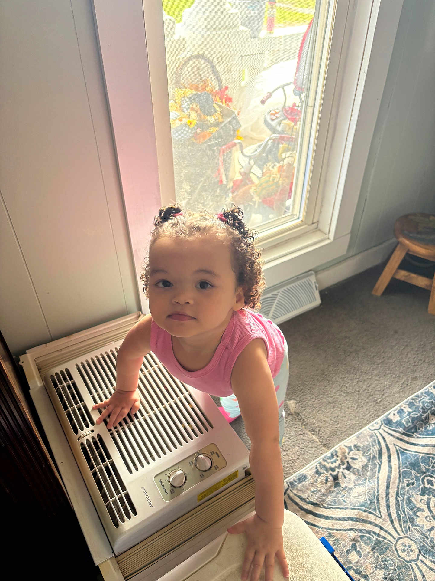 Maddilynn is registered to the contest to win money with this photo: toddler, child, indoor, curly_hair, pink_shirt, window, air_conditioner, carpet, stool, home, light, hands, curiosity, person, portrait, standing, floor, furniture, hair_ties, expression