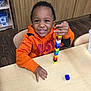 Adrian is registered to the contest to win money with this photo: child, smiling, orange_hoodie, table, wooden_chair, colorful_blocks, building_blocks, indoor, play, happy, furniture, wood_floor, cabinet, education, preschool, person, toy, learning, young_child, stacking