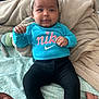baby, headband, blue_shirt, nike, black_pants, bed, blanket, patterned_bedspread, infant, person, indoors, skin, face, hand, foot, smile, cute, clothing, rest, cozy
