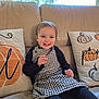 toddler, child, smiling, sitting, couch, pillows, pumpkin, window, indoor, clothing, dress, black_sleeves, happy, face, hair, home, cozy, natural_light, cute, portrait