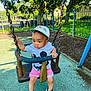 toddler, child, swing, playground, cap, shorts, tshirt, mickey_mouse, shoes, outdoor, trees, fence, sunlight, shade, play, park, metal_chains, rubber_surface, summer, daytime