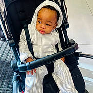 Jesse is registered to the contest to win money with this photo: baby, child, stroller, onesie, striped_clothing, infant, black_hair, seated, indoors, floor, curious_expression, hand, footwear, safety_strap, metal_frame, fabric, person, casual_wear, baby_clothes, toddler
