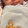 baby, newborn, sleeping, smiling, portrait, face, indoors, blanket, knitted_blanket, swaddled, cozy, teddy_bear, bear, embroidery, blue_bow, clothing, infant, close_up, peaceful, vertical_photo
