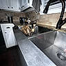cat, tortoiseshell_cat, pet, animal, sink, kitchen, faucet, cabinet, countertop, backsplash, dish_towel, indoors, home, plumbing, stainless_steel, sitting, looking_at_camera, flash_photography, domestic_scene, funny_pet
