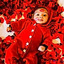 baby, infant, child, red_onesie, knit_outfit, rose_petals, lipstick_marks, face, big_eyes, surprised_expression, lying_on_back, cozy_blanket, buttons, soft_texture, indoor, portrait, close_up, playful, floral, red_color