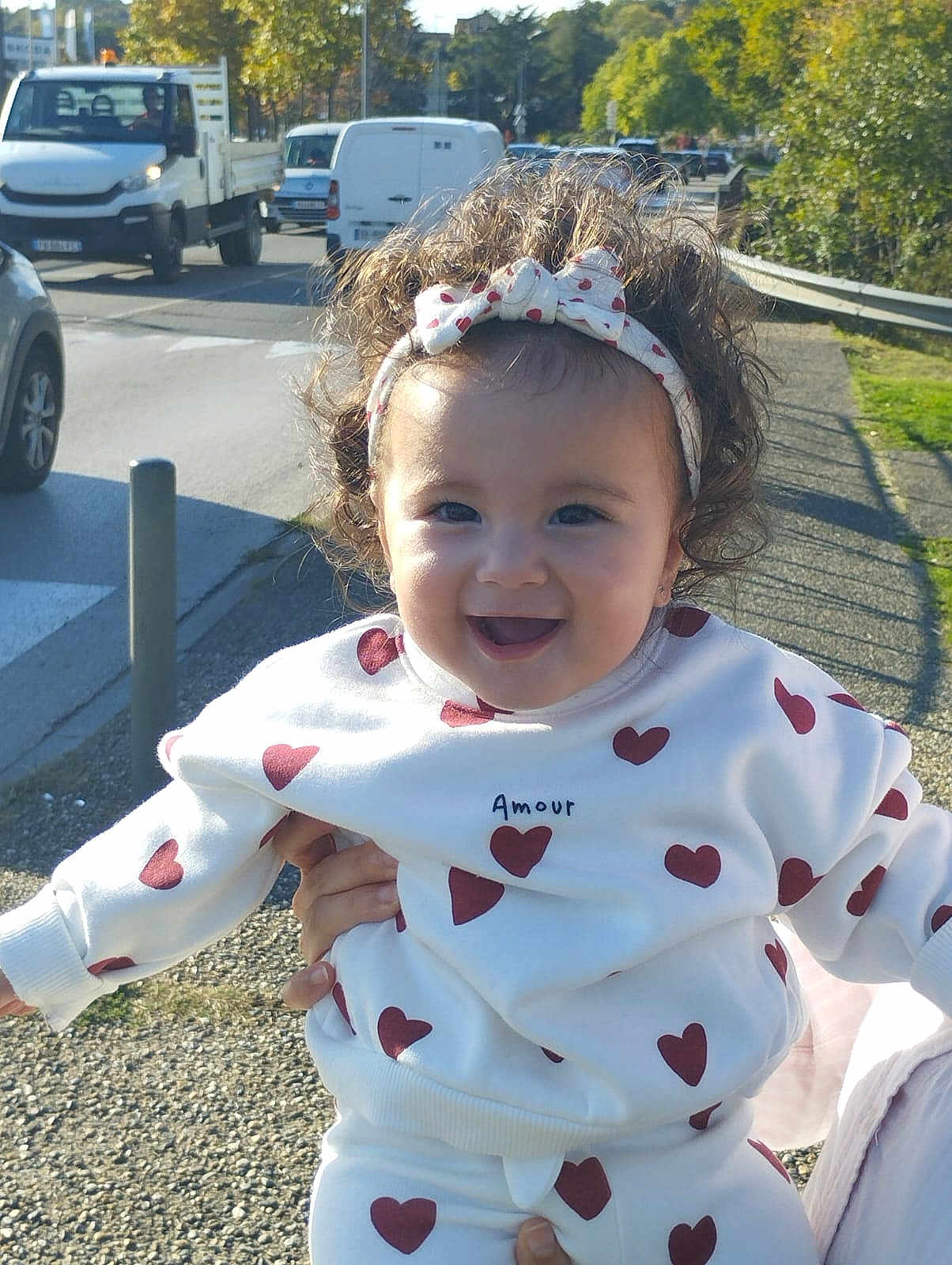 Aryam a rejoint le concours — aidez-le/la à gagner de superbes lots ! toddler, child, curly_hair, headband, heart_pattern, white_clothing, outdoor, sunlight, smile, happy, person, holding_hands, road, vehicles, trees, sidewalk, daytime, casual_wear, cute, playful
