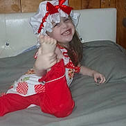 Delilah is registered to the contest to win money with this photo: child, smiling, red_bow, bonnet, red_pants, white_top, apple_pattern, foot, hand, blanket, wooden_wall, happy, playful, indoors, person, cute, young, cheerful, posing, casual