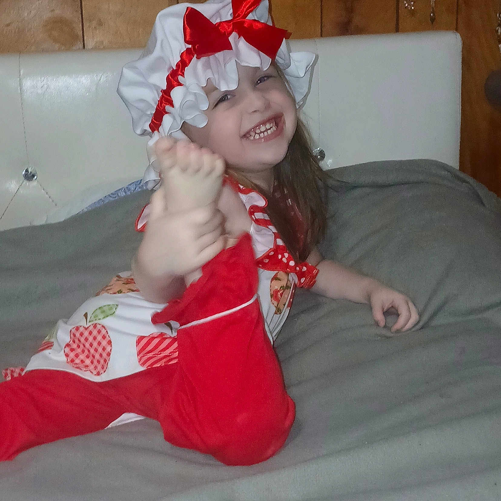 Delilah is registered to the contest to win money with this photo: child, smiling, red_bow, bonnet, red_pants, white_top, apple_pattern, foot, hand, blanket, wooden_wall, happy, playful, indoors, person, cute, young, cheerful, posing, casual
