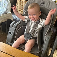 Sam is registered to the contest to win money with this photo: baby, high_chair, child, smile, arms_up, striped_shirt, indoor, happy, seatbelt, wooden_table, furniture, home, person, infant, face, legs, playful, cute, safe, expression