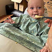 Sam joined the competition — help win amazing prizes! baby, feeding, highchair, spoon, bib, dinosaur_pattern, messy, food, happy, smiling, indoor, table, hand, person, child, face, blue_eyes, clothing, home, couch