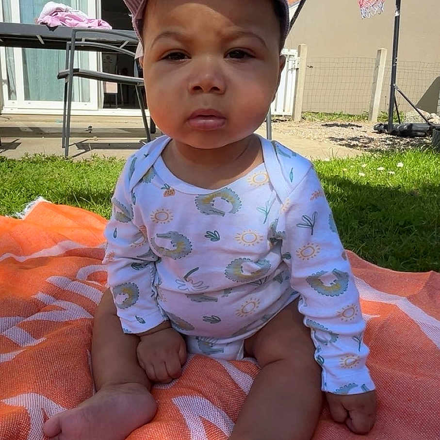 Soren participe au concours pour gagner de l'argent avec cette photo : baby, child, outdoor, grass, blanket, cap, onesie, sunlight, basketball_hoop, garden, fence, patio, table, chair, person, foot, hand, face, expression, summer